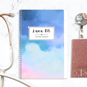 May include: A spiral-bound notebook with a blue and pink watercolor sky design featuring the text Luna RN POCKETBUDDY, a white stethoscope, and a glittery pink badge holder with a small unicorn and heart charm attached to a grey reel.