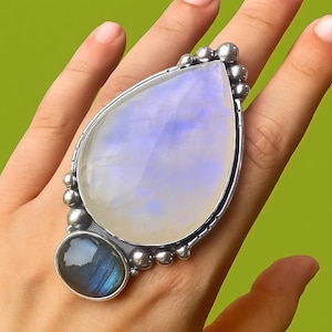 Puede incluir: Un anillo grande, plateado, con una piedra lunar iridiscente en forma de pera y una labradorita ovalada más pequeña. La piedra lunar es de color blanco lechoso y azul pálido. El anillo se muestra en una mano sobre un fondo verde.