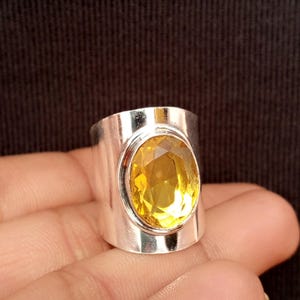 Könnte beinhalten: Ein breiter Silberring mit einem großen, oval geschliffenen, facettierten gelben Citrin, der in einer glatten, polierten Silberfassung sitzt. Die Metalloberfläche ist reflektierend und schlicht, was den Kontrast zwischen dem gelben Stein und Silber betont.
