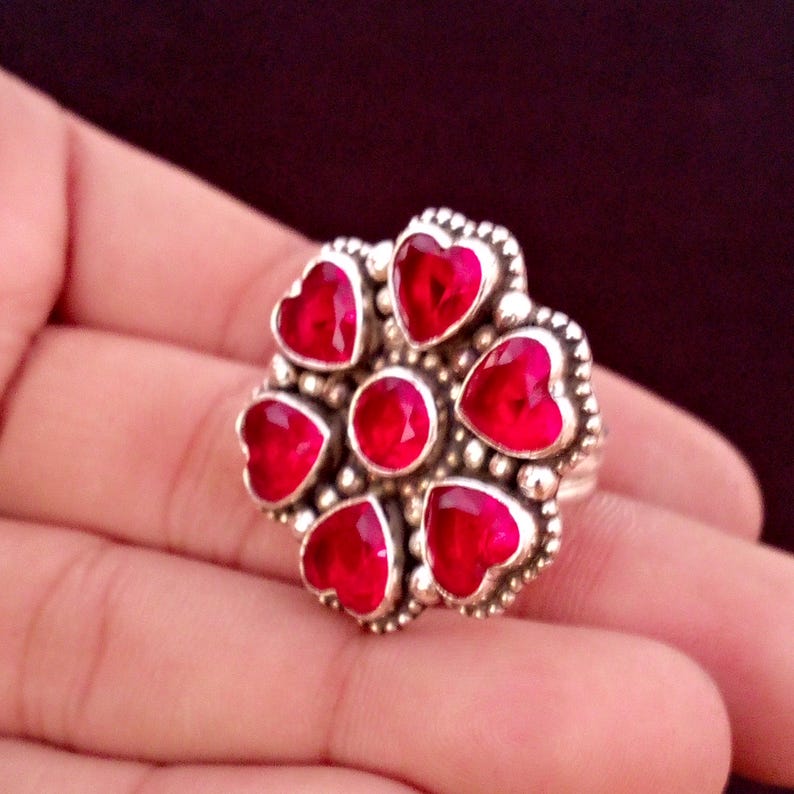 May include: A silver ring featuring a flower design with heart-shaped red gemstones. The ring has a central round gemstone and is surrounded by six heart-shaped stones. The ring is held in a hand.