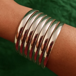 Può includere: Un ampio bracciale a fascia composto da sette singoli anelli in metallo color argento, lisci e lucidi, disposti fianco a fianco per creare un aspetto sovrapposto. La superficie di ogni anello è altamente riflettente.