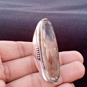 Può includere: Un anello grande, di forma ovale, con una pietra preziosa di quarzo fumé sfaccettata. L'anello ha una fascia color argento con un design dettagliato e strutturato attorno alla pietra preziosa. La pietra preziosa è di colore marrone chiaro.
