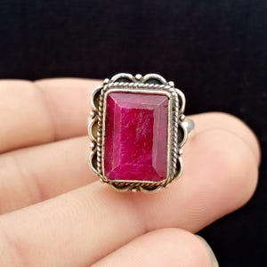 May include: A silver ring featuring a large, rectangular, deep red gemstone. The gemstone is set in a decorative, ornate silver frame with a rope-like border. The ring is shown against a dark background.