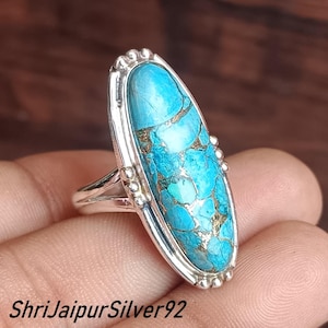 May include: A silver ring with an oval turquoise stone. The stone has a blue and gold pattern. The ring has a decorative silver border. The text "Shri Jaipur Silver92" is at the bottom of the image.