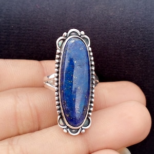 May include: A silver ring featuring a large, oval lapis lazuli stone. The deep blue stone has flecks of gold and is set in a decorative silver frame with a beaded border. The ring's design is ornate and eye-catching.