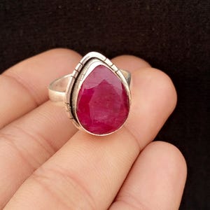 Puede incluir: Un anillo de plata con una gran piedra preciosa roja oscura en forma de lágrima. La piedra preciosa está engastada en una banda de plata detallada con detalles grabados. El anillo se muestra sobre un fondo oscuro.