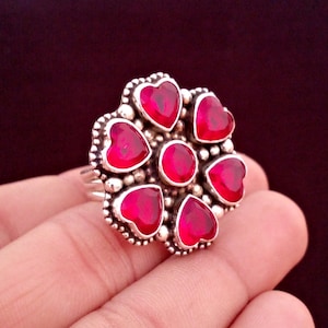 May include: A silver ring featuring a floral design with heart-shaped, deep red gemstones. The ring's setting has a beaded border, and a central round stone. The ring is displayed on a hand.