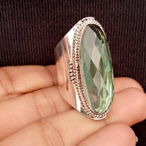 Puede incluir: Un anillo grande, plateado, con una piedra preciosa verde pálido ovalada y facetada. El anillo tiene una banda gruesa y detalles decorativos en forma de cuerda alrededor de la piedra. La piedra preciosa mide unos 3,8 cm de largo.