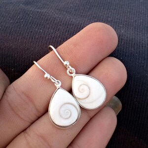 Puede incluir: Pendientes colgantes de plata con colgantes blancos en forma de lágrima. Cada colgante presenta un diseño en espiral. Los pendientes se muestran sobre un fondo oscuro.