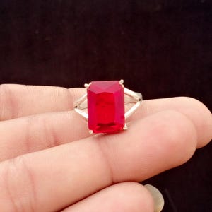 May include: A silver ring featuring a large, rectangular, faceted red gemstone set in a four-prong mount. The band of the ring splits into two separate silver arms as it reaches the setting, creating an open, geometric design on both sides.