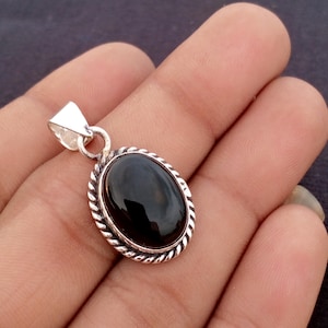 May include: Oval black gemstone pendant with a silver-colored metal setting. The pendant has a rope-like border and a triangular bail. The gemstone is smooth and polished, reflecting light. The pendant is held in a hand.
