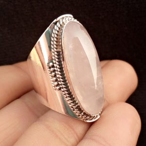 May include: A silver ring featuring a large, oval rose quartz gemstone. The gemstone is a soft pink color and is set in a silver band with a braided rope design around the stone. The ring has a wide band.