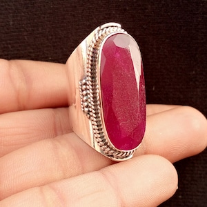May include: A silver ring with a large, oval-cut ruby gemstone. The ruby is a deep red color and is set in a wide band with decorative silver detailing. The ring is held in a hand against a dark background.