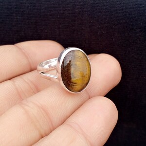 May include: A silver ring featuring an oval tiger's eye gemstone. The gemstone has a golden-brown color with streaks. The ring's band has a split design. The ring is displayed against a dark background.