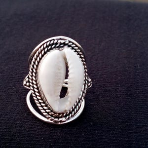 Pode incluir: Um anel de tom prateado com uma concha de búzios branca no centro. A concha está envolvida por uma borda de metal com design de corda trançada que forma uma moldura oval. O aro do anel é visível sob a cravação decorativa polida.