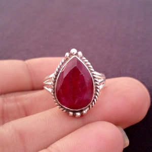 May include: A silver ring featuring a large, teardrop-shaped, faceted ruby gemstone. The ruby is a deep red color and is set in a silver band with a twisted rope design around the stone. The ring has a simple, elegant design.