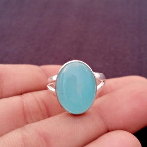 May include: A silver ring featuring a large, oval-shaped, light blue gemstone. The gemstone is set in a simple, polished silver band with a split shank design. The ring is displayed against a purple background.