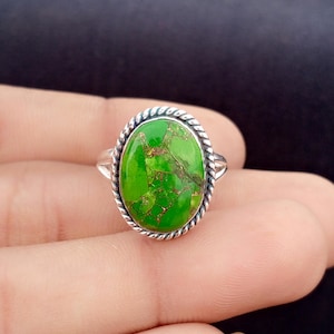 May include: Oval-shaped silver ring featuring a vibrant green stone with brown veining. The stone is set within a twisted rope-like border. The ring is displayed against a neutral background, highlighting the jewelry.