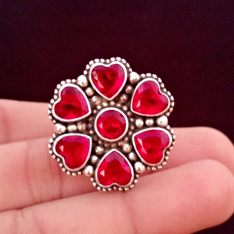 May include: A silver-toned ring featuring a flower design with six heart-shaped red gemstones surrounding a central round red gemstone. The ring has a beaded border and is set against a dark background.