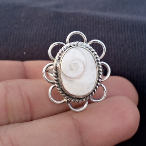 Op de afbeelding: Een zilveren ring met een ovale, witte steen met een spiraalpatroon. De steen is gezet in een gedraaide, touwachtige rand en omgeven door een bloemvormige zetting. De ring wordt in een hand gehouden.