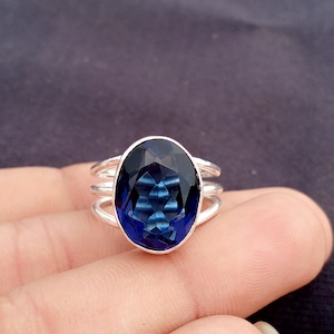 May include: A silver ring with a large, oval-shaped, deep blue gemstone. The gemstone has a faceted surface that reflects light. The ring has a triple-band design. The ring is a piece of jewellery. The ring is a piece of jewellery.