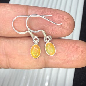May include: A pair of silver drop earrings featuring small, oval-shaped opals with iridescent orange and yellow flashes. The stones are set in simple silver bezels, suspended from curved silver ear wires with a coiled wire detail at the top.