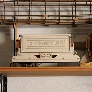 May include: A wooden model of a Chevrolet truck bed, painted in a light cream color. The word "CHEVROLET" is displayed prominently on the tailgate. The Chevrolet logo is visible below the tailgate. The model is displayed on a wooden shelf.