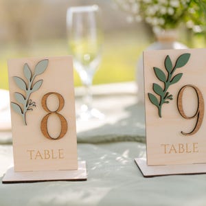 Peut inclure: Deux numéros de table en bois avec des accents de feuilles vertes. Les numéros sont 8 et 9. Les pancartes indiquent "Table".