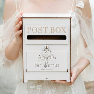Puede incluir: Un buzón de madera blanco con las palabras "POST BOX" en una fuente marrón claro. La caja tiene una ranura para el correo y un panel inferior con los nombres "Amelia & Benjamin" y la fecha "APRIL 23TH 2028". Un candado en forma de corazón asegura la caja.