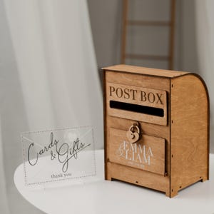 Op de afbeelding: Een houten postbus met de woorden "POST BOX" op de voorkant. Onder de gleuf zit een deur met een hartvormig slot en de namen "EMMA & LIAM" en de datum "APRIL 21TH 2023". Een helder bord met "Cards & Gifts thank you" staat links.