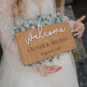 Puede incluir: Un letrero de boda de acrílico transparente con un panel de madera. El panel de madera tiene la palabra "welcome" en escritura blanca, junto con los nombres "OLIVER & MELISSA" y la fecha "12 de agosto de 2025". El letrero está decorado con hojas verdes de eucalipto.