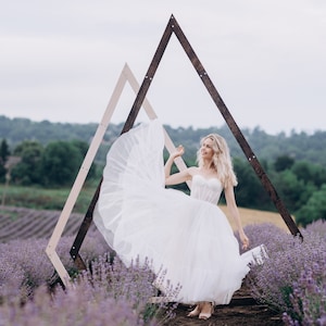 Puede incluir: Una novia con un vestido de novia blanco se encuentra en un campo de lavanda, sonriendo y sosteniendo su vestido. Está de pie frente a un arco de madera.