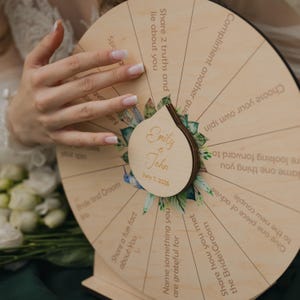 May include: A wooden wedding game wheel with prompts like "Share a fun fact about you" and "Share how you met the Bride/Groom." The wheel is engraved with "Emily & John" and the date "July 7, 2025."