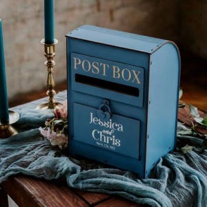 May include: A blue wooden post box with the words "POST BOX" in gold lettering. Below the slot, a panel displays "Jessica and Chris" and the date "March 27th, 2023". The box has a heart-shaped lock and is set on a wooden surface.