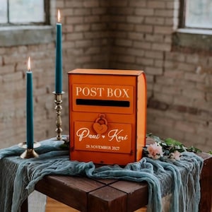 Może przedstawiać: Pomarańczowa metalowa skrzynka pocztowa z białym napisem "POST BOX". Poniżej widnieją imiona "Paul • Keri" i data "28 listopada 2027". Dwa turkusowe świeczniki w mosiężnych świecznikach flankują skrzynkę, ustawioną na drewnianym stole z niebieską tkaniną.