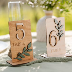 May include: Two wooden table number signs with a rustic finish. The number 5 is on a brown sign with a green leafy branch. The number 6 is on a light brown sign with a green leafy branch. Both signs have the word "TABLE" printed below the number.