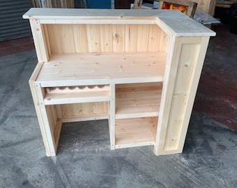 May include: A light-colored, L-shaped wooden bar with a countertop, shelving, and a wine rack. The bar is constructed from unfinished wood, showcasing the natural grain and texture. It features a built-in wine rack and multiple shelves for storage.