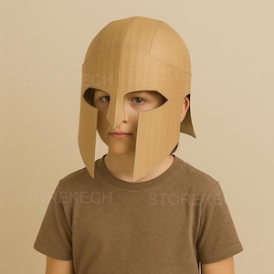 May include: A cardboard Spartan helmet with eye openings and cheek guards. The helmet is tan and appears to be handmade. The person in the image is wearing a brown t-shirt. The helmet is a costume piece.