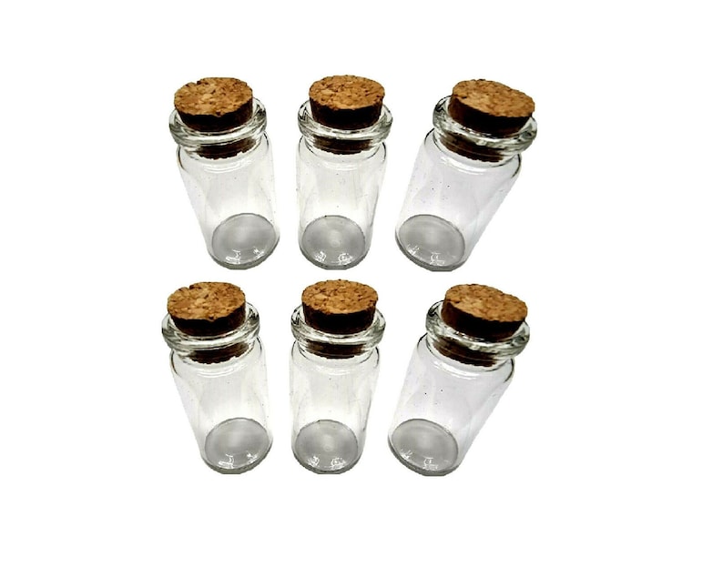 May include: Six small, clear glass bottles with cork stoppers. The bottles are cylindrical with rounded bottoms. The corks are a light brown color. The bottles are arranged in a slightly angled pattern against a white background.