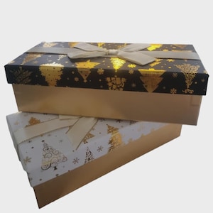 May include: Two rectangular gift boxes, one atop the other. The top box has a black lid with gold Christmas tree and snowflake patterns, and a gold base. The bottom box has a white lid with gold Christmas tree patterns and a gold base. Both have cream-colored ribbon bows.