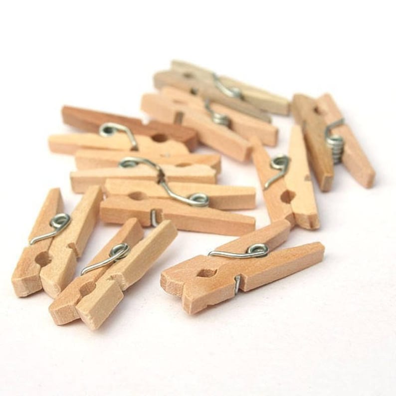 May include: A group of wooden clothespins with metal springs. The clothespins are a light brown color and are arranged in a pile.