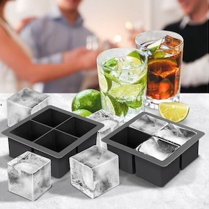 May include: Two black silicone ice cube trays with four compartments each. The trays are filled with clear ice cubes. There are also three loose ice cubes on a white surface. Two glasses with ice and drinks are in the background.