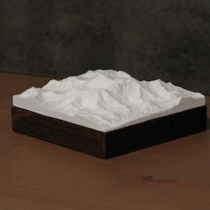 Op de afbeelding: Een wit gipsmodel van een bergketen, geplaatst op een donkerhouten basis. Het model is een gedetailleerde weergave van een berglandschap.