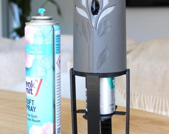 May include: A light blue aerosol air freshener can with floral graphics next to a black plastic stand holding a gray cylindrical cover featuring a cutout leaf pattern. The air freshener can has the text "Denk mit" and "Duft Spray" printed on it.