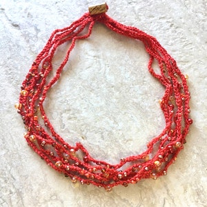May include: A handmade red necklace with multiple strands. The necklace is made of woven red thread and features small, round, amber-colored beads. The necklace is displayed on a light gray surface.