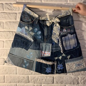 Könnte beinhalten: Ein Jeansrock mit Patchwork-Design, bestehend aus verschiedenen blauen und weißen Stoffen, Spitzenbesatz und gestickten Blumenmotiven. Der Rock hat ein weiß-blaues Blumenband, das um die Taille gebunden ist.