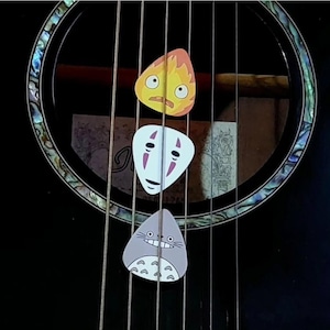 May include: Three guitar plectrums featuring characters from Studio Ghibli films. The picks are on the strings of a black acoustic guitar. The top pick is a yellow and orange flame, the middle is a white mask, and the bottom is a grey Totoro.
