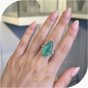 May include: A large, pear-shaped, emerald-green gemstone ring. The ring has a silver band with small accent stones. The gemstone is the focal point, showcasing its clarity and color. The ring is displayed on a hand.