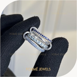 May include: A multi-band ring featuring three interconnected silver-toned bands. One band is set with dark blue stones, one with light blue stones, and one with clear stones. The text FORME JEWELS is visible in the image, layered over the rings.