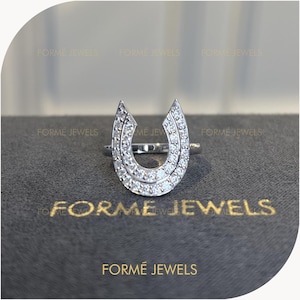 May include: A silver-toned ring featuring a horseshoe-shaped centerpiece adorned with numerous small, clear, sparkling stones. The text FORME JEWELS is visible multiple times in the background and printed in gold on the dark surface below the ring.